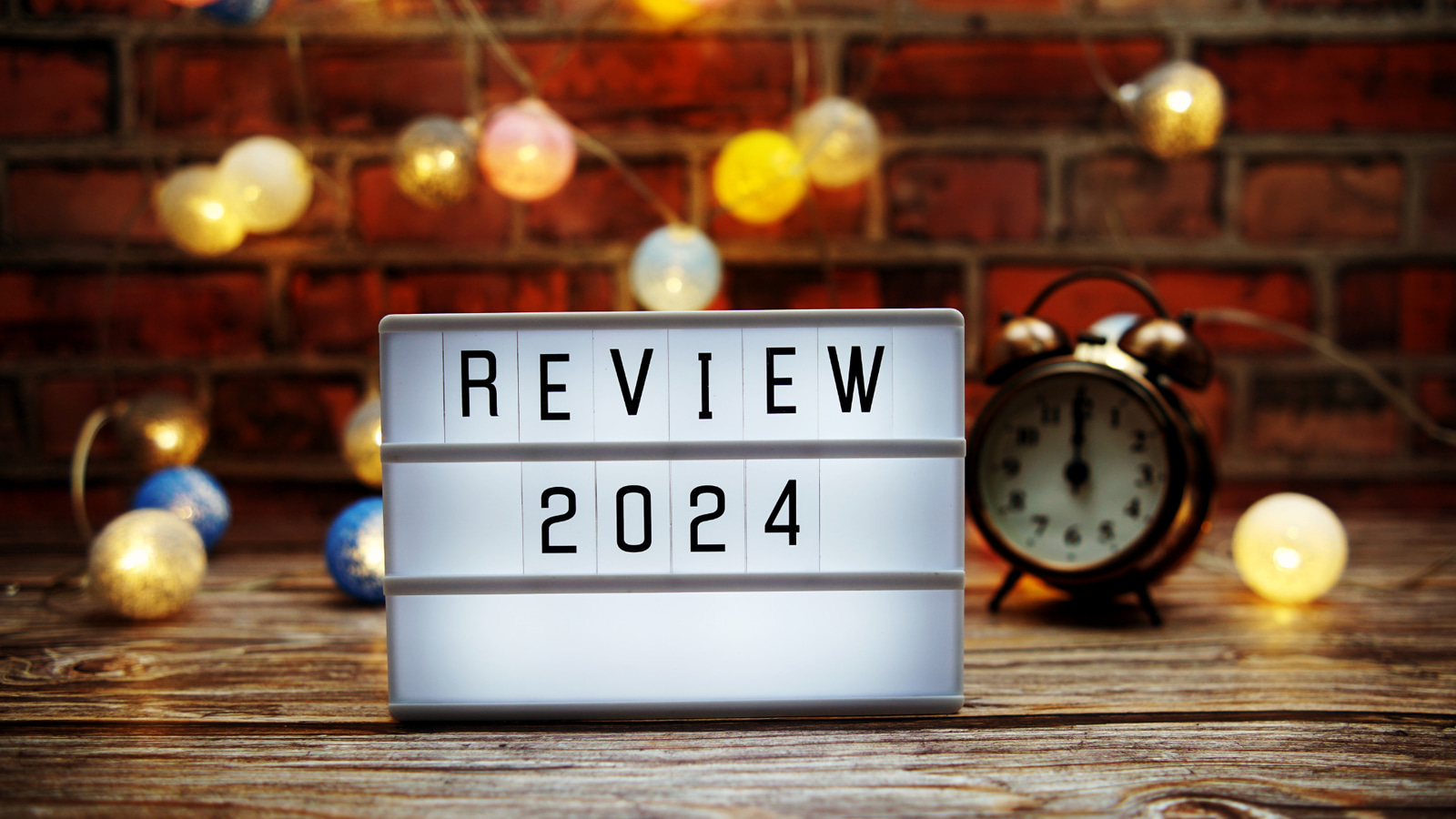 Foto einer Leuchttafel mit der Aufschrift: Review 2024, im Hintergrund sind kleine Licher und einen Wecker der Mitternacht zeigt