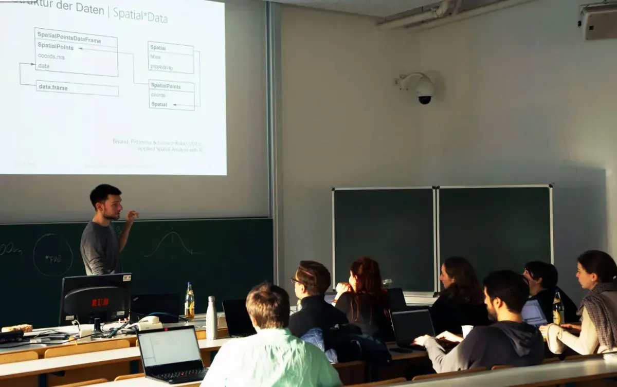 Bild eines Seminarraums. Eine Person steht vorne und erklärt einer Gruppe von ca. 10 sitzenden Personen etwas. Im Hintergrund die an die Leinwand geworfenen Folien. Alle Personen haben einen Laptop vor sich. 