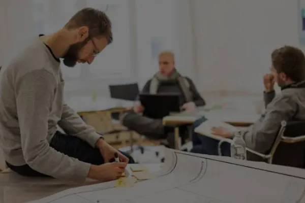 Eine Person mit braunen Haaren und Vollbart sitzt auf einem Tisch und schreibt auf Post-It Karten. im Hintergrund sitzen zwei weitere Personen und unterhalten sich. Eine Person hat einen Laptop auf dem Schoß. 